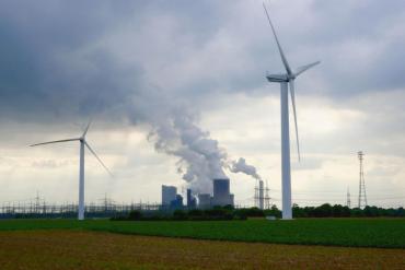 Photo with wind turbines in the foreground with factory building spewing emissions and transmission grid in the background