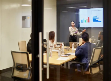 Photo of conference room with workers seated at a table with a woman presenting and a graph on the projection screen.