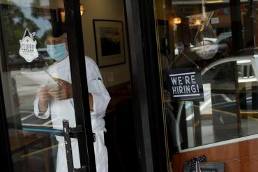 A worker at a restaurant in a mask with a sign in the window that says 'We're Hiring'.