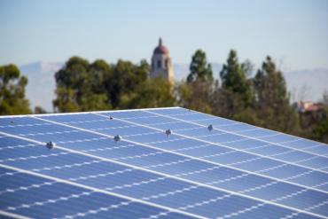 Photo of solar panels on a rooftop at Stanford with the Hoove tower, trees and mountains in the background