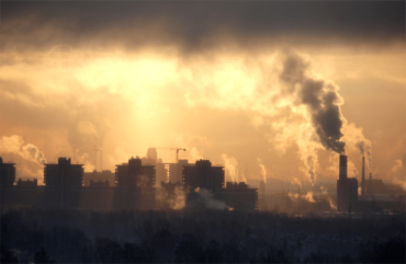 A Photo of a city in the distance with smoke stacks with smoke filling a yellow and dark sky