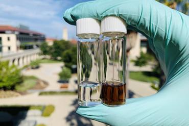 Gloved hand in the foreground holding two test tubes one with a clear liquid, the other with a brown liquid against the backdrop of the School of Engineering quad