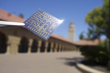 An array of artificial synapses being held in foreground with tweezers against a backdrop of the Stanford quad.