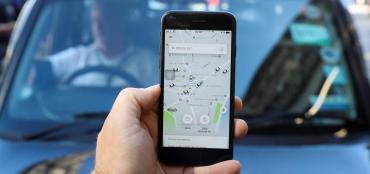 Image of a hand holding a cellphone against a backdrop of a driver in a vehicle